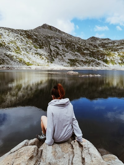 白天，一个穿着灰色连帽衫的女人坐在湖边的岩石上